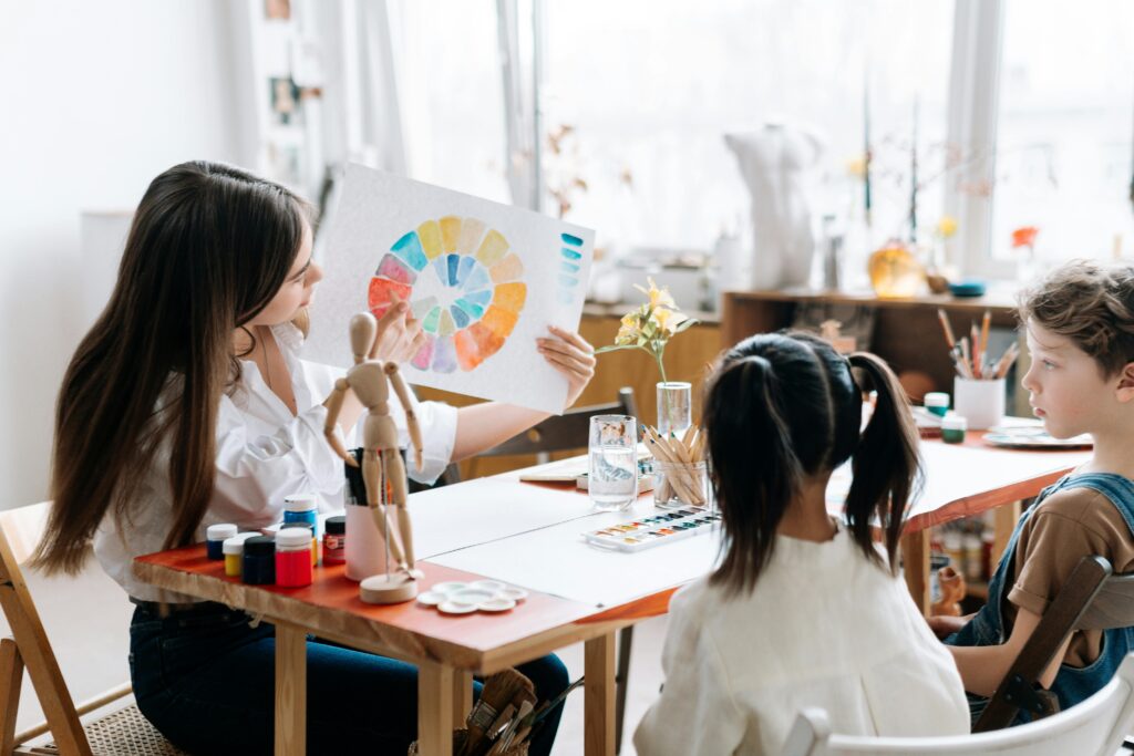 Woman holding up a watercolor image of the color wheel to show children seated at a table with painting supplies.