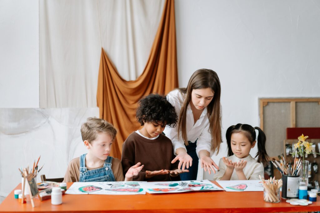 Children seated at a table painting. Woman stands at table instructing the children.