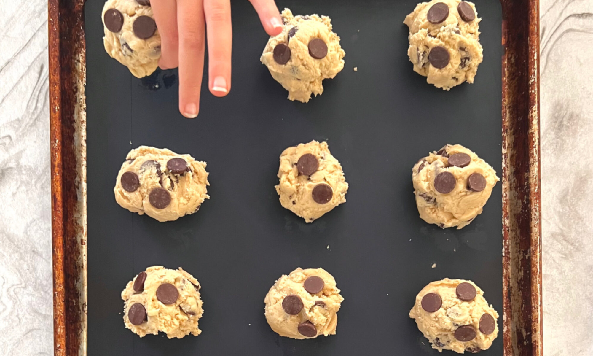 Homeschool Science Activity: Chocolate Chip Cookie Chemistry - Balls of Chocolate Chip Cookie Dough on Baking Pan with Silicone Mat Child Hand is Placing Last Ball of Cookie Dough