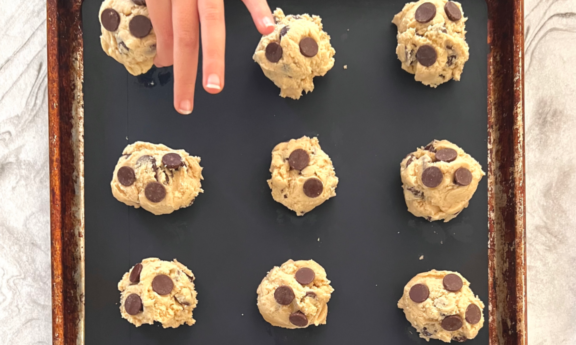 Homeschool Science Activity: Chocolate Chip Cookie Chemistry - Balls of Chocolate Chip Cookie Dough on Baking Pan with Silicone Mat Child Hand is Placing Last Ball of Cookie Dough