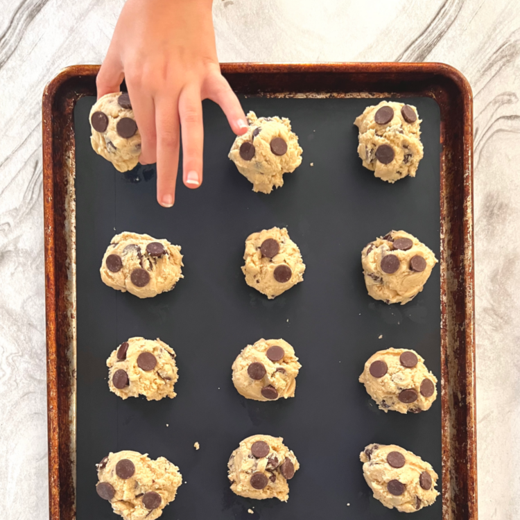 Homeschool Science Activity: Chocolate Chip Cookie Chemistry - Balls of Chocolate Chip Cookie Dough on Baking Pan with Silicone Mat Child Hand is Placing Last Ball of Cookie Dough