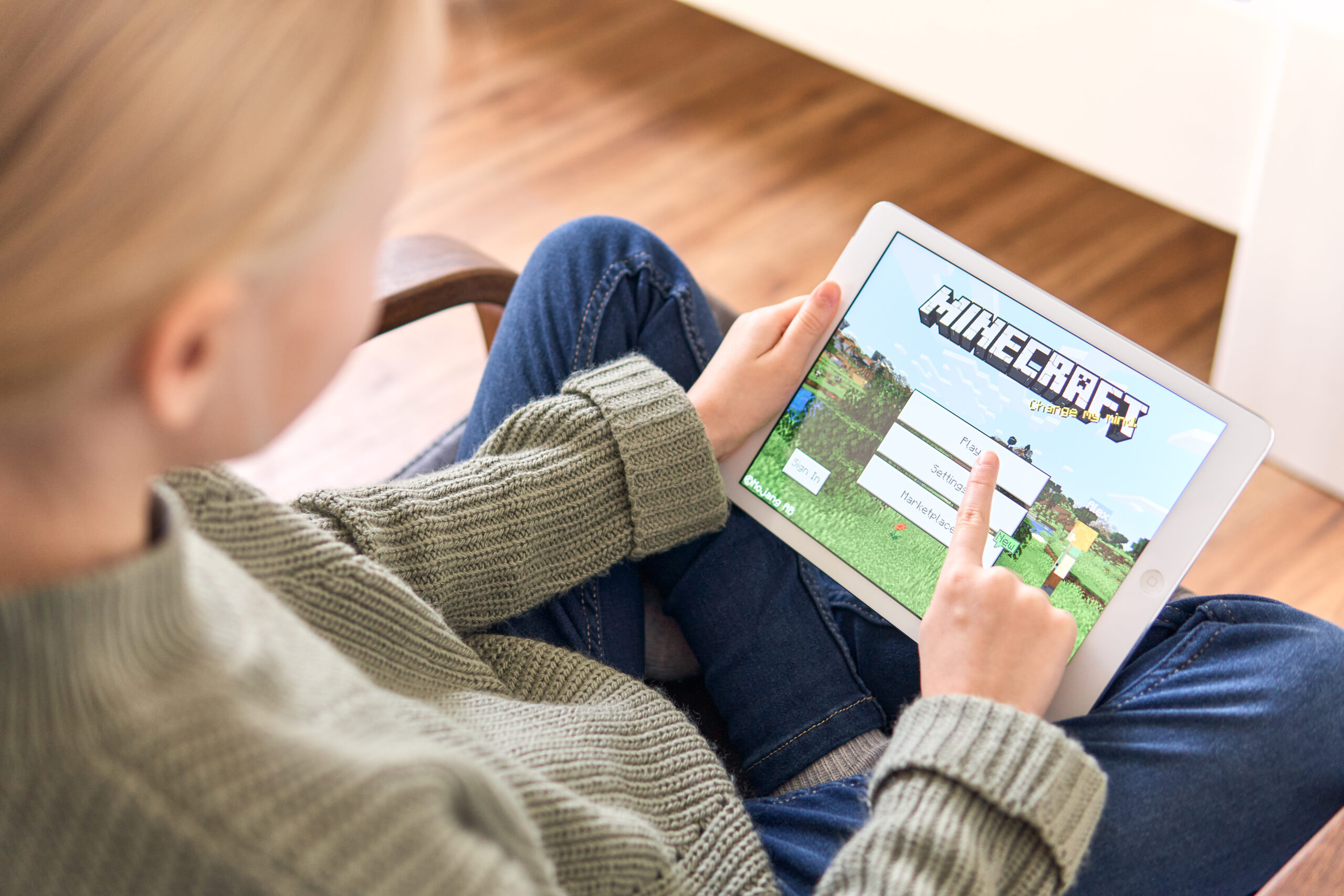 Child with blonde hair sits holding an iPad with the intro screen to Minecraft showing